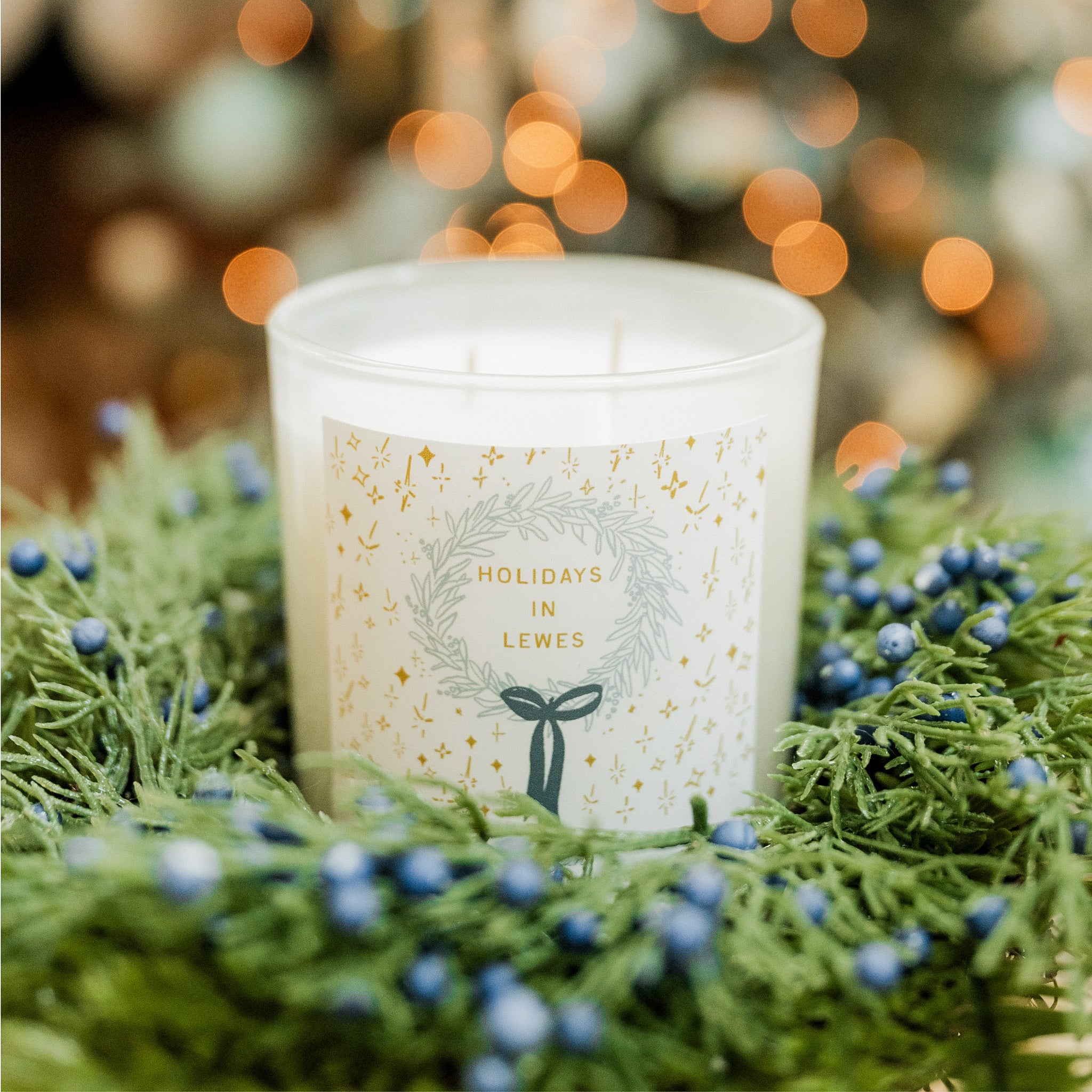 Candle labeled 'Holidays in Lewes' surrounded by greenery and berries with a blurred festive background.