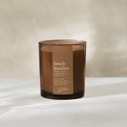 A beach bonfire scented candle in a translucent amber glass jar with a brown paper label, set against a warm beige background.