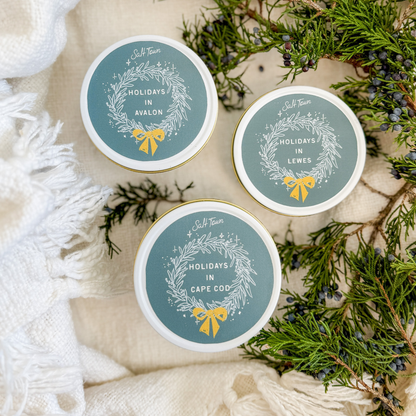 Three small containers with festive labels on a decorative background with greenery and white fabric.